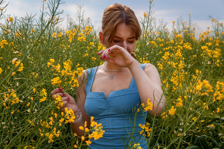 Woman in blue dress standing among blooming yellow wildflowers, sneezing and covering her face, possibly due to seasonal allergiesの写真素材
