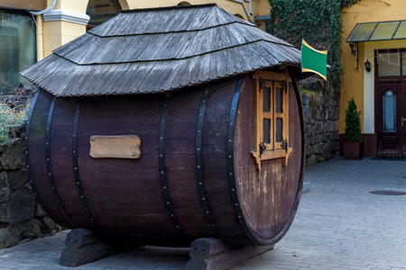 Quirky wooden structure designed like a giant barrel with a small window, decorative metal hinges, and a shingled roof, placed in a courtyard near a traditional buildingの写真素材