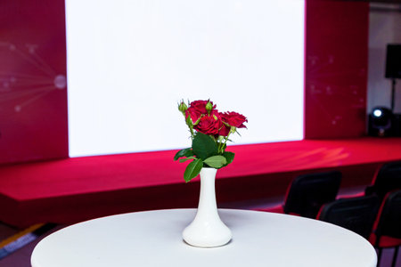 Small white vase with red roses placed on a round table in front of a red stage and projection screen in a conference or presentation settingの写真素材