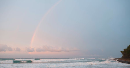 Rainbow, pink colors of clouds and sea foam on waves. Dark forest on shore. Dramatic colors from frontal lighting sunset.の写真素材