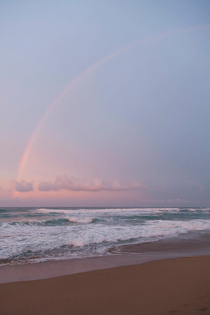 Rainbow, pink colors of clouds and sea foam on waves. Big surf waves.の写真素材