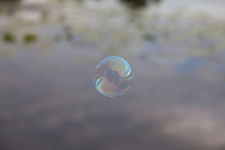 Rainbow soap bubble on the background reflections in the river. The sky with clouds and aquatic plants.の写真素材
