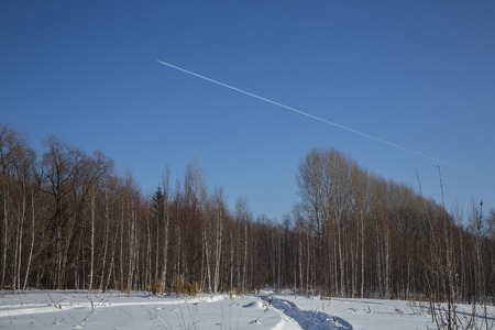 the trail of the plane in the blue sky against the background of the winter forest. path in the snow. Trees.の写真素材