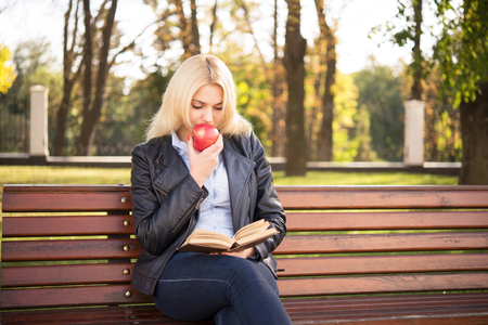 beautiful blond girl with an apple sitting on a bench and reading a bookの写真素材
