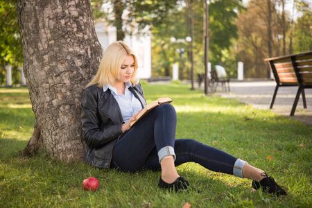 beautiful blond girl reading a book and sitting under a treeの写真素材