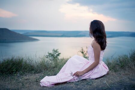 beautiful girl in a dress sitting on the bank of the river and watching the sunsetの写真素材