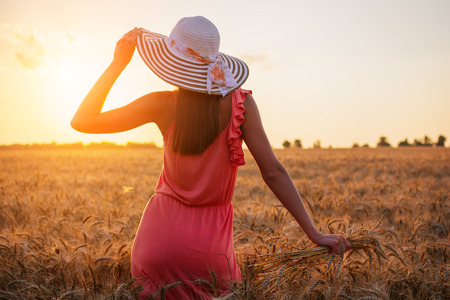 Beautiful young woman with brown hear wearing rose dress and hat enjoying outdoors looking to the sun on perfect wheat field on sunsetの写真素材