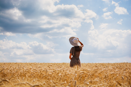 Beautiful young woman with brown hear wearing rose dress and hat enjoying outdoors looking to the horizont on perfect wheat fieldの写真素材