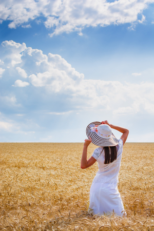 Beautiful young woman with brown hear wearing rose dress and hat enjoying outdoors looking to the horizont on perfect wheat fieldの写真素材