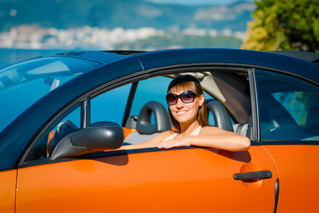 Beautiful young woman with long hair sitting and smiling in orange cabriolet at the Mediterranean sea coastの写真素材