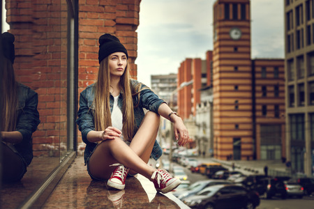 Outdoor lifestyle portrait of pretty young sitting girl, wearing in hipster swag grunge style urban background. Retro vintage toned image, film simulation.の写真素材