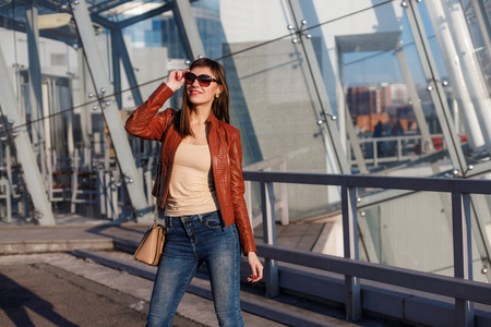 Portrait of beautiful brunette young woman in nice red brown jacket, denim jeans and sunglasses. Genuine leather bag, high heels shoes. Spring autumn fashion trendy photo of lady on urban background.の写真素材