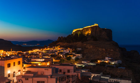 Scenic landscape photo of night Lindos town and castle on Rhodes island, Dodecanese, Greece. Panorama with bright lights, mountains and sea. Famous tourist destination in South Europeの写真素材