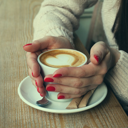 Close-up female hands holding cup with coffee cappuccino with foam with pattern heart. Perfect red gel polish manicure. wood natural table.の写真素材