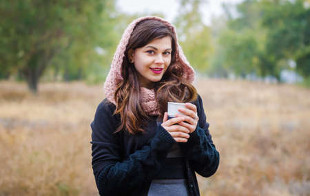 Young smiling girl in a scarf and hat with a cup of coffee (tea) in the hands of autumn outdoors.