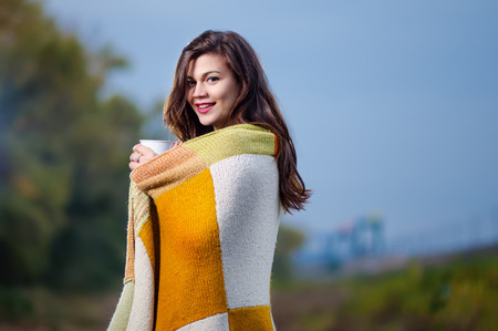 Young cheerful girl with a blanket on his shoulders and a cup of coffee (tea) in his hands. Autumn eveningの写真素材