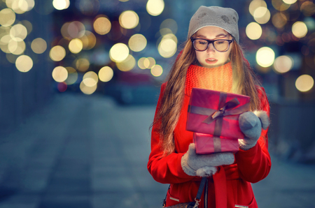 Attractive young girl with long hair in a red coat and scarf with the gift in a box with the shining rays on a beautiful evening background with bokeh.の写真素材