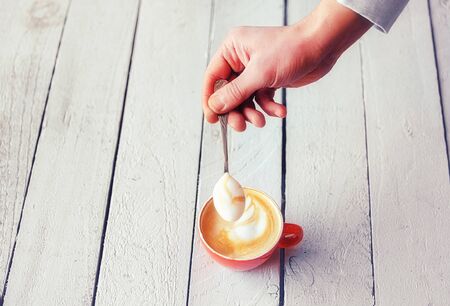 Barista's hand holding a spoon with a delicious cream of a coffee latte with pattern on a ceramic red cup on the white wooden table.の写真素材