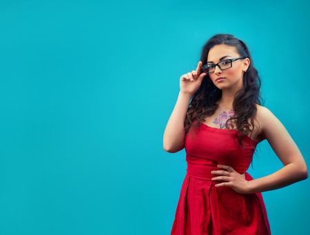 Young serious girl with makeup, wearing glasses and a red dress on a blue backgroundの写真素材