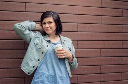 Smiling brunnete girl with tunnels in the ears in a beige  leather jacket  with a cup of coffee...