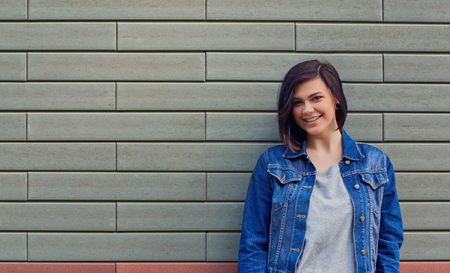 Beautiful smiling young woman in a blue jeans jacket near a textured brick wall.の写真素材