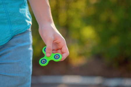 Teenager hand holding  trendy relaxation gadget fidget spinner. Man on the azure shirt and blue jeans playing with green spinner outdoors  on the bright floral bokeh. Shallow DOF.の写真素材