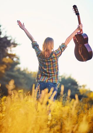 Photo from the back of a happy girl with her hands up with an acoustic guitar on a autumn meadow in sunset light.の写真素材