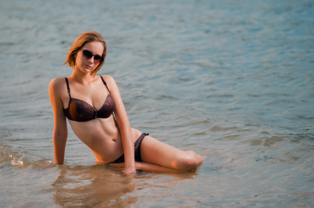 Pensive sexy  caucasian young woman in a glasses on the beach, is sitting on azure water. Recreation and pampering on the seashore (ocean, river, lake) in sunset light.の写真素材