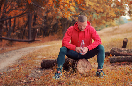 young athletic man in a red sports jacket with a hood sits on a log and holds bottle with water after running on a red autumn forest.の写真素材