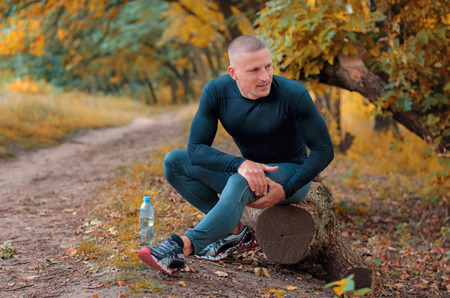 A young athletic  jogger in black tight fitting sportswears and  sneakers sits on a log, is in pain and holding a hip with his hands after cramping on a autumnall forest background.の写真素材