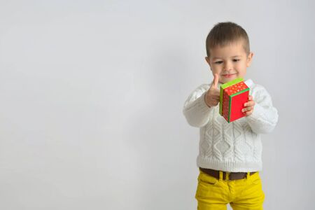 A happy smiling  boy in a white sweater and yellow jeans is shows a  good gesture on  hand with thumb up. Child looking of a red cardboard box. Isolate on white background. Christmas conceptual photo.の写真素材