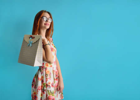 Portrait of a caucasian teenager  in glasses in an  white dress. Girl holding in the hand stylish carton shopping bag. Conceptual studio photo of a young  model  on the blue background.の写真素材