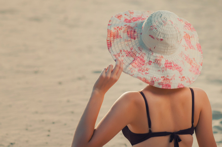 Tall caucasian slaender girl holds a hand over the edge of the cap on the beach, is sitting on golden sand. Recreation and pampering on the seashore (ocean, river, lake) in summer and sunny day.の写真素材