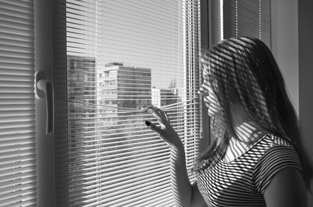 the girl looks out the window through the blindsの写真素材
