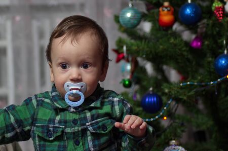 A little boy in a green shirt with a pacifier stands at the holiday tree.の写真素材