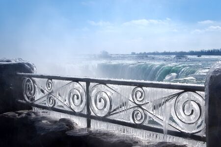 Decorative iron fence covered by thick layer of frozen mist. Niagara Falls on background.の写真素材