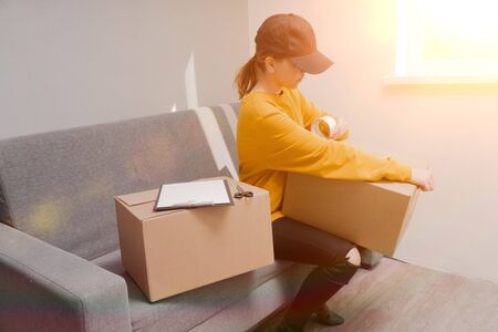 young courier girl seals a cardboard box with duct tape on a light backgroundの写真素材