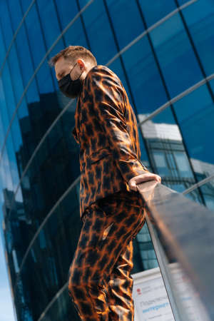 Portrait of a young man in a medical black mask looks forward against the background of a business center. average plan of a businessman in a business suit.の写真素材