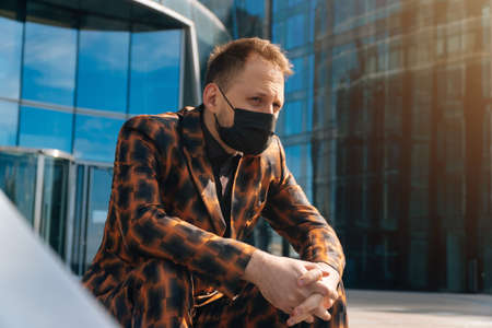 Portrait of a sad young man in a medical mask looking down ahead against the background of a business center. average plan businessman is sitting in a business suit.の写真素材