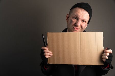 Young sad beggar man holds a felt-tip pen to write text on cardboard. Homeless Assistance Concept.