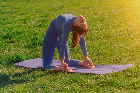 A young caucasian girl is standing in a pose on a mat on green grass during sunset. The concept of using physical exercises for balance of mind. Fitness Stretching Trainingの写真素材