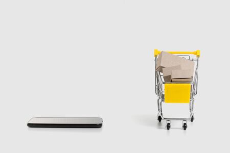 A shopping cart in an online store next to a smartphone on one white background. Paper boxes in a construction trolley.の写真素材