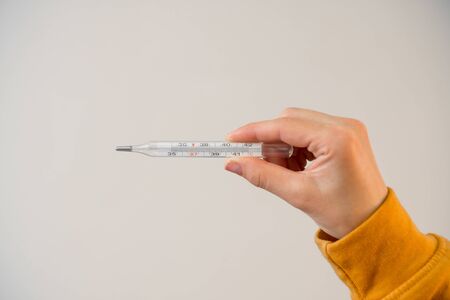 Thermometer in a female hand with simple manicure on a background of a light wall. Take temperature...