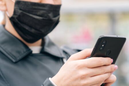 Close-up women's hands hold a phone against the background of a face in a medical mask. The concept of safety is about your health. Girl calls taxiの写真素材