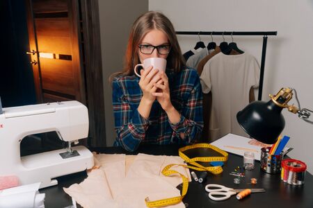 A young girl a seamstress designer with glasses holds a cup with a hot drink in her hands and looks at the camera. Lunch break concept.の写真素材
