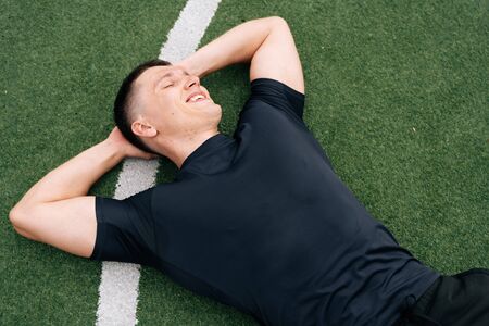 A portrait of a smiling sports man lies on the green grass on a football field. The concept of fatigue from outdoor trainingの写真素材