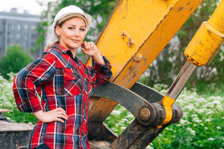 Woman in construction clothes with a white helmet in a plaid shirt on the background of an excavator.の写真素材