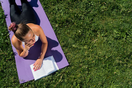 A brooding young girl with a book view from above, a sporty woman lies on a yoga mat on a summer dayの写真素材