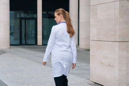 A young girl doctor in a white robe goes against the background of the medical clinicの写真素材