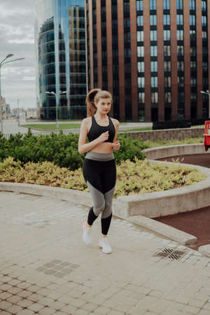A young blonde sports girl in a black suit runs along modern houses